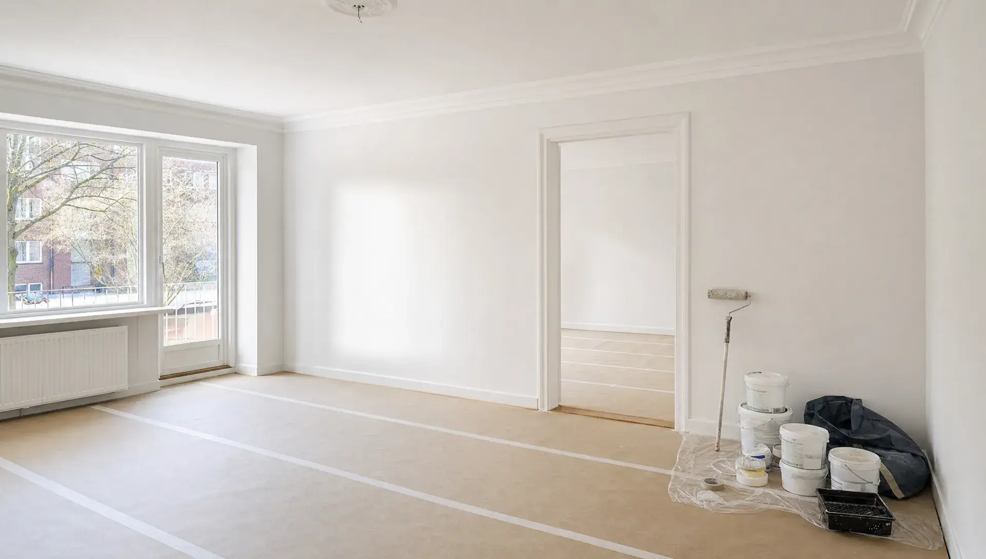 En tom lejlighedsstue med afdækkede gulve, nymalede vægge og loft samt malerudstyr samlet ved en væg, så rummet fremstår klart til indflytning eller opfriskning.