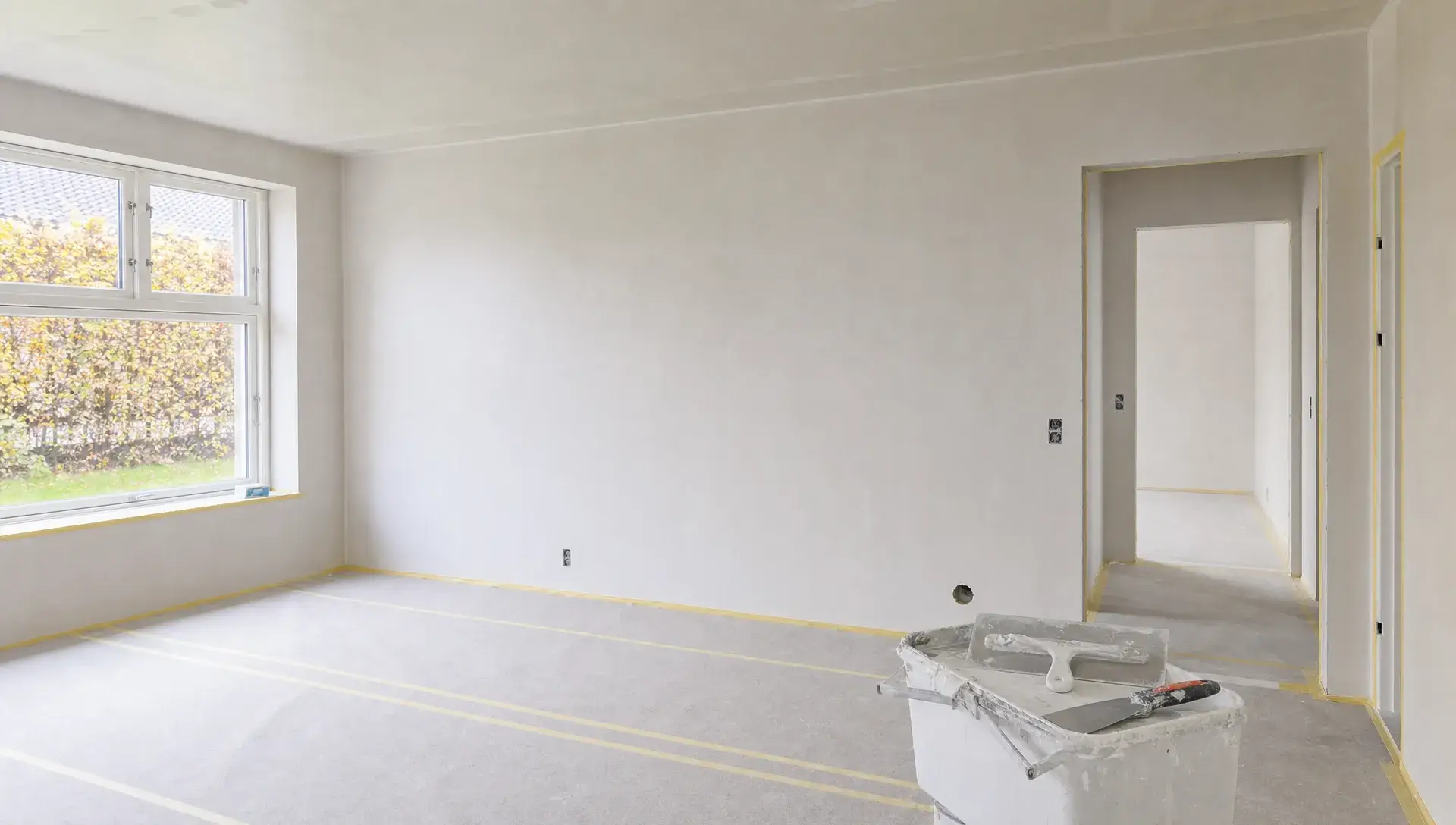 Et lyst rum under renovering med glatte fuldspartlede vægge, afdækket gulv og spartelværktøj klar til næste behandling.