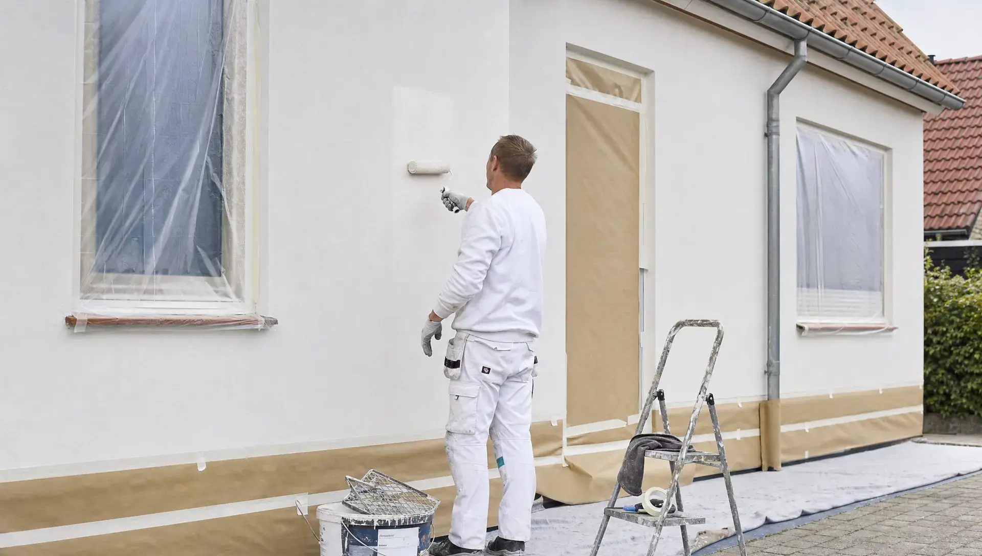 En maler arbejder på en klargjort facade på et dansk hus med værktøj og afdækning omkring muren.
