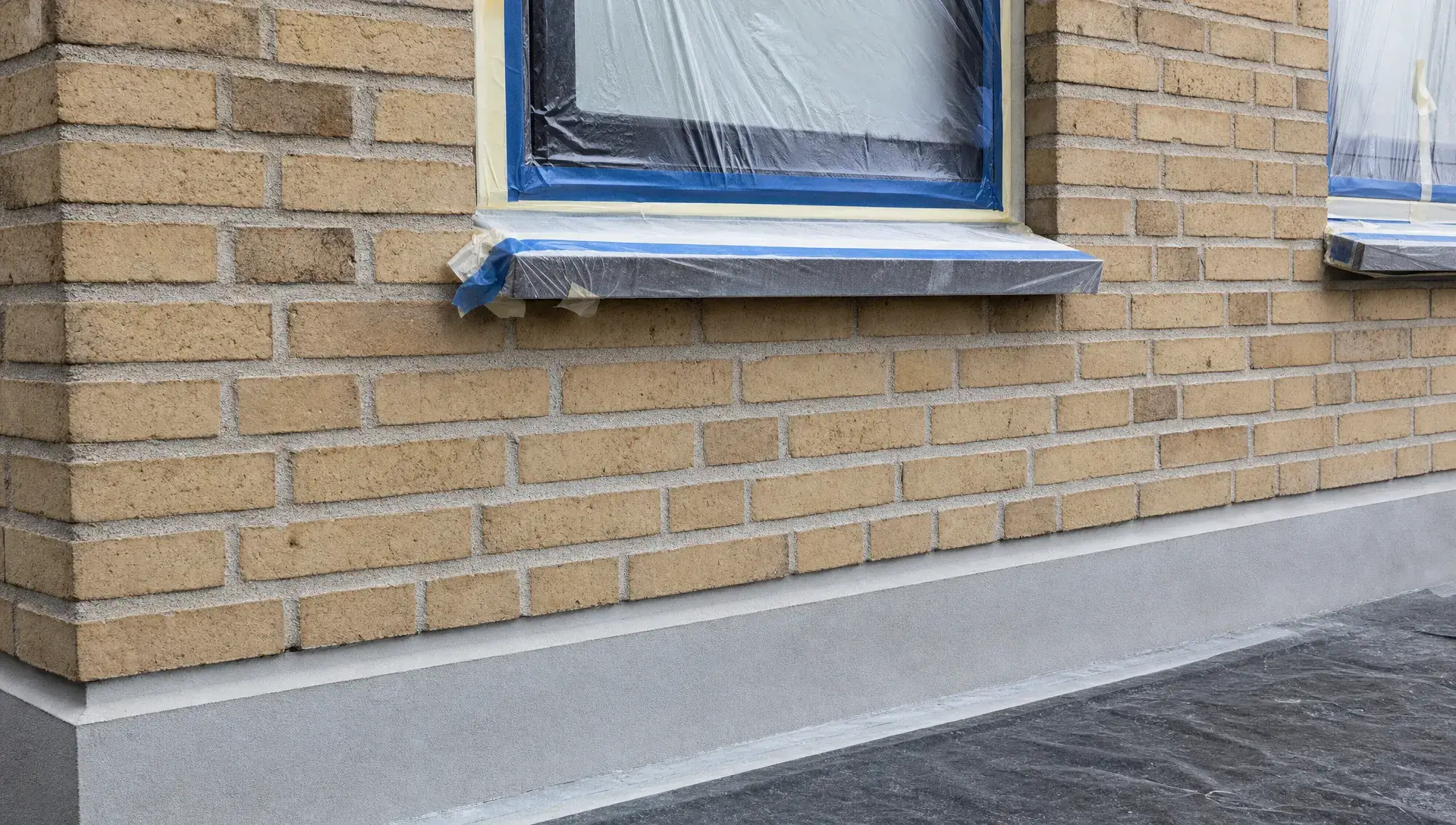 Et nærmere udsnit af en murstensfacade under klargøring, hvor afdækning ved vinduer, en ren sokkel og præcist arbejde omkring fuger og murværk viser forarbejdet til en ensartet facadefinish.