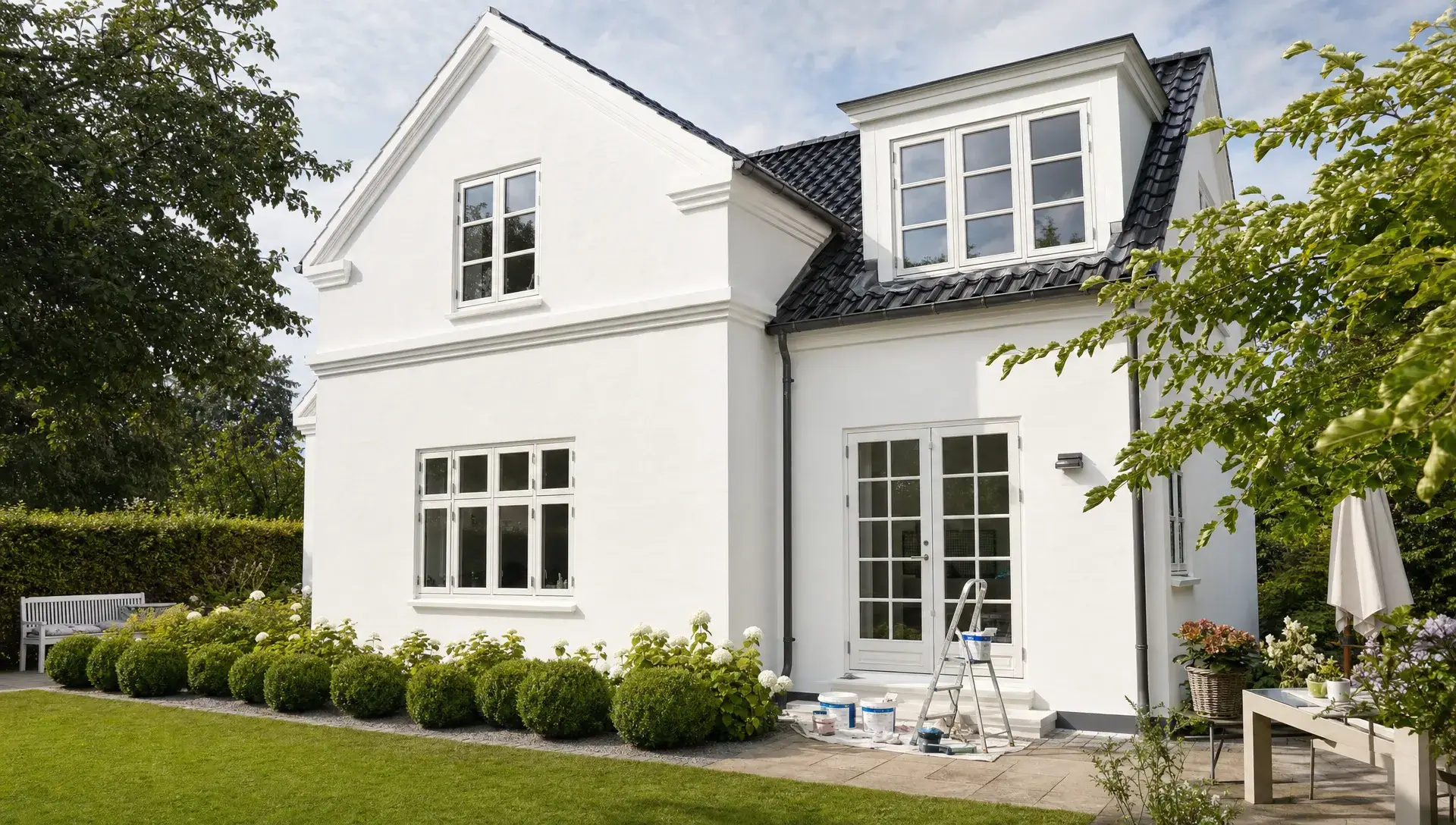 Nymalet lys facade på en villa i Nordsjælland set fra haven med rene linjer, velholdt murværk, grøn beplantning foran huset og diskret malerudstyr ved bygningen.